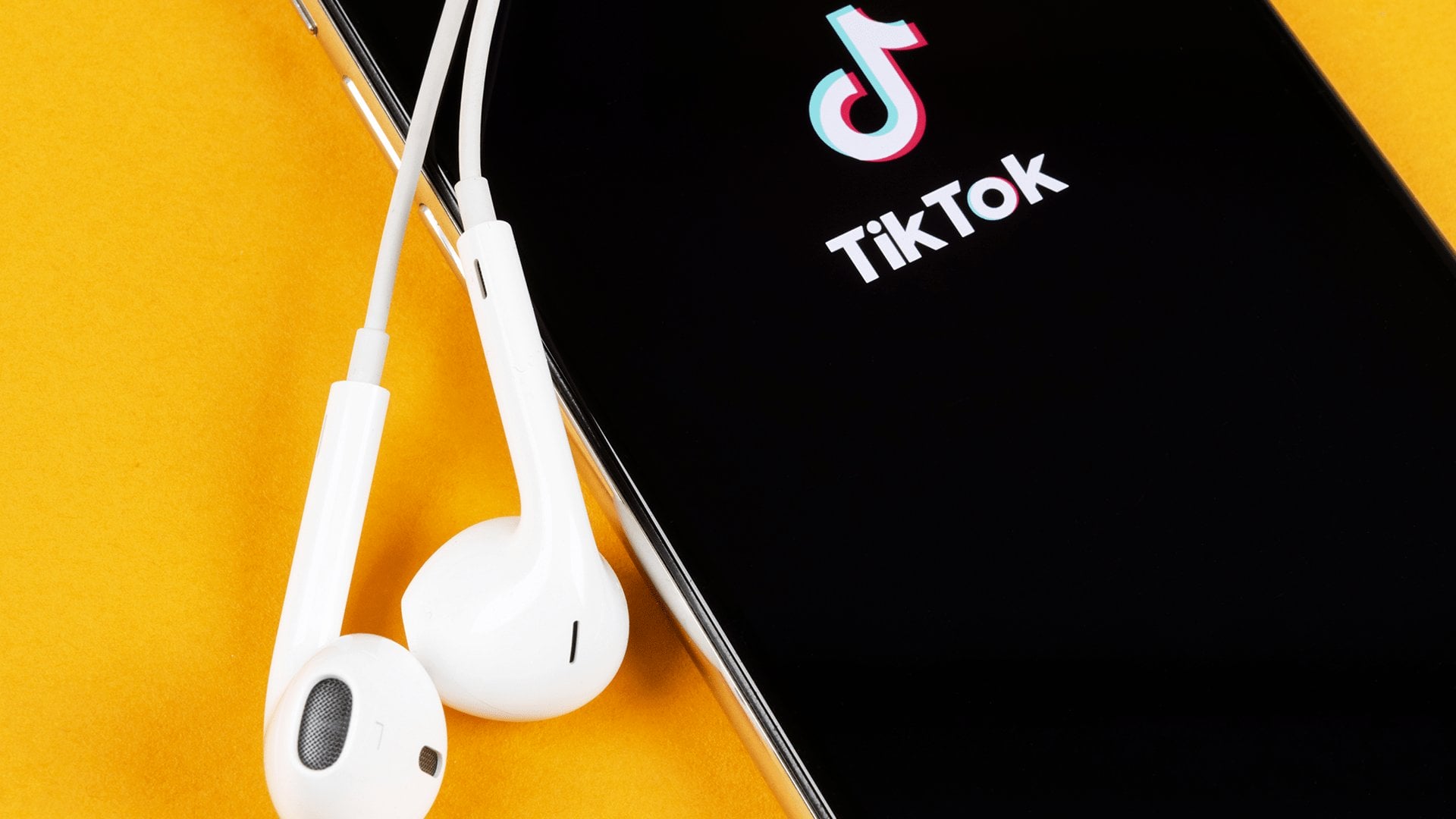 Een smartphone met het TikTok-app-logo op het scherm bevindt zich op een levendig geel oppervlak, met een paar witte bedrade oortelefoons bovenop het apparaat. Ondernemers op TikTok gebruiken dit platform om innovatieve ideeën te delen en in contact te komen met een breder publiek.