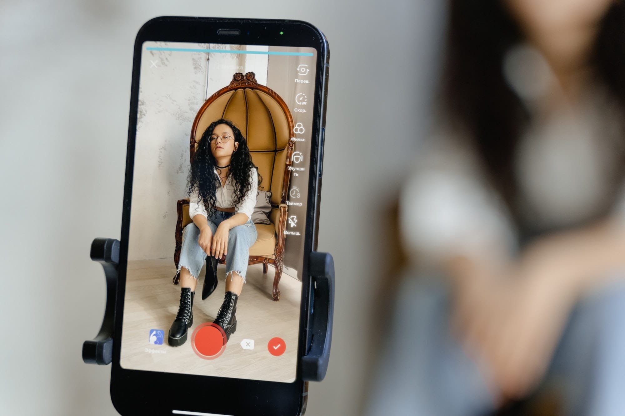 Een smartphone op een statief maakt een TikTok voor je bedrijf-video van een persoon die op een grote vintage stoel zit, zichtbaar via het telefoonscherm. De persoon draagt een wit overhemd, een gescheurde spijkerbroek en zwarte laarzen, met lang krullend haar. De achtergrond is wazig.