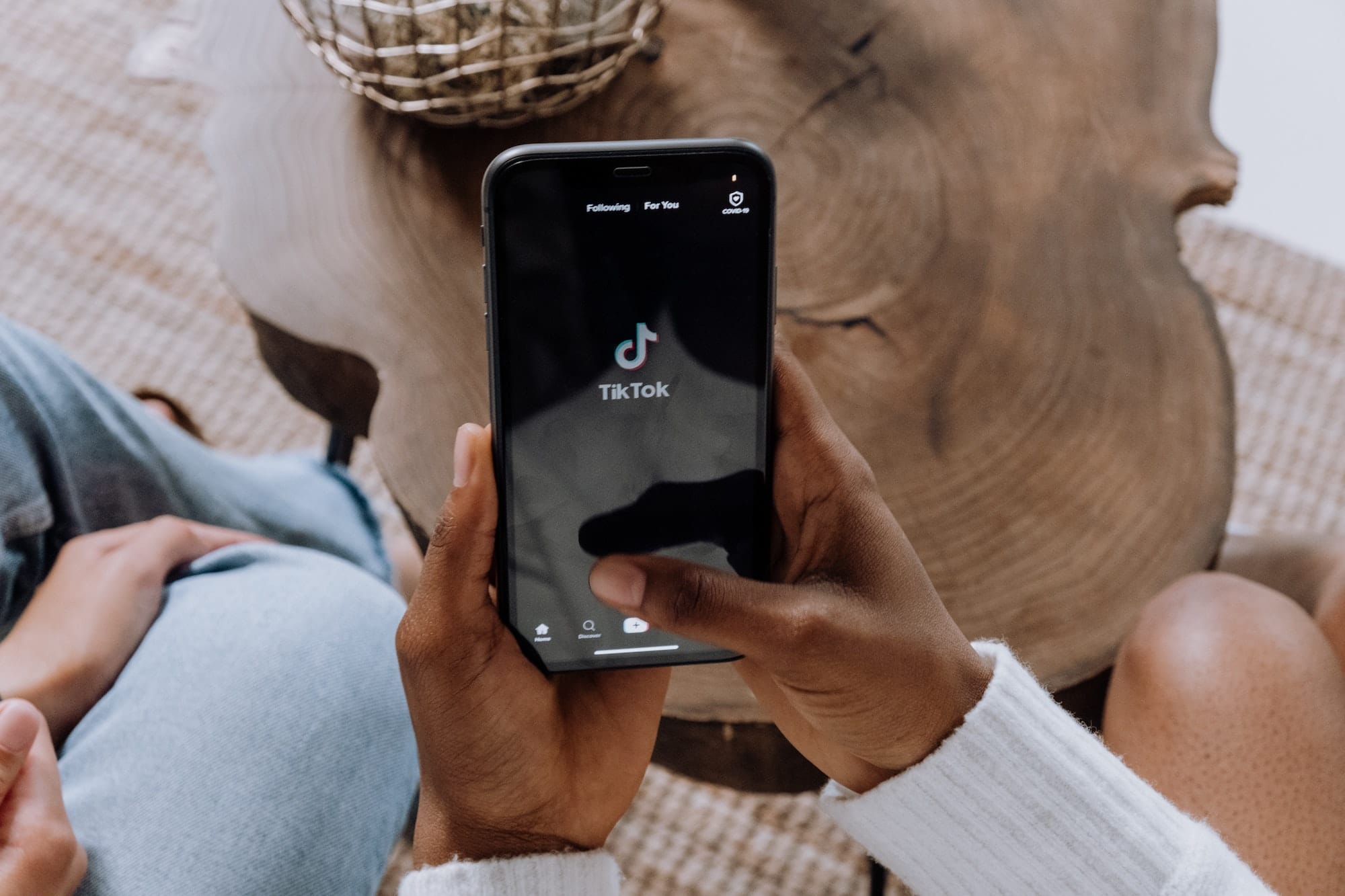 Close-up van twee handen die een smartphone vasthouden waarop de TikTok-app op het scherm wordt weergegeven boven een houten salontafel. Op de achtergrond staat een decoratieve geweven kom. Het lijkt erop dat de persoon bezig is met het gebruik van TikTok voor je bedrijf.