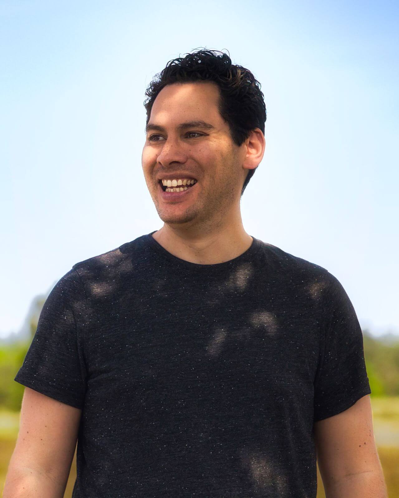 Portret van een lachende Bram van Houtum in een zwart T-shirt, buiten gefotografeerd met een blauwe lucht op de achtergrond.