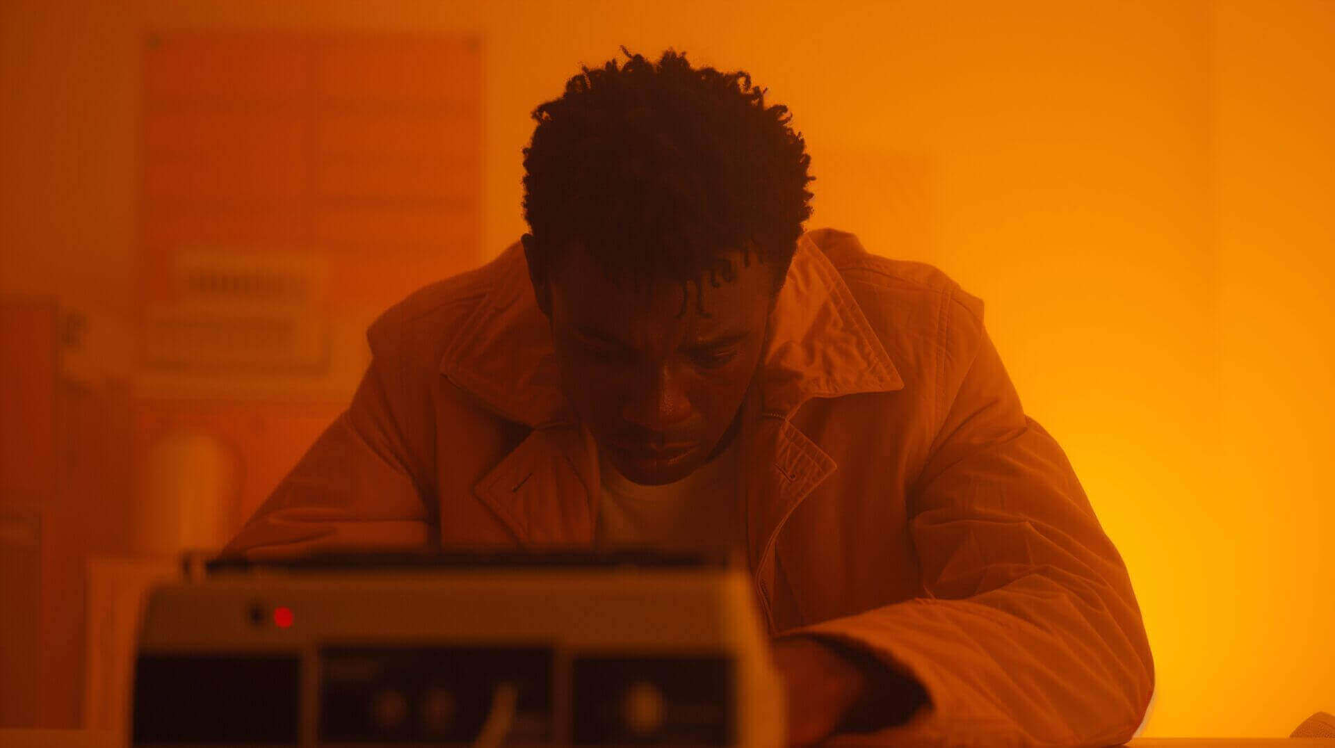 Een man met kort krullend haar in een beige jas zit aan een tafel en kijkt naar beneden, omgeven door een oranje gloed. De achtergrond is wazig met enkele apparaten zichtbaar.