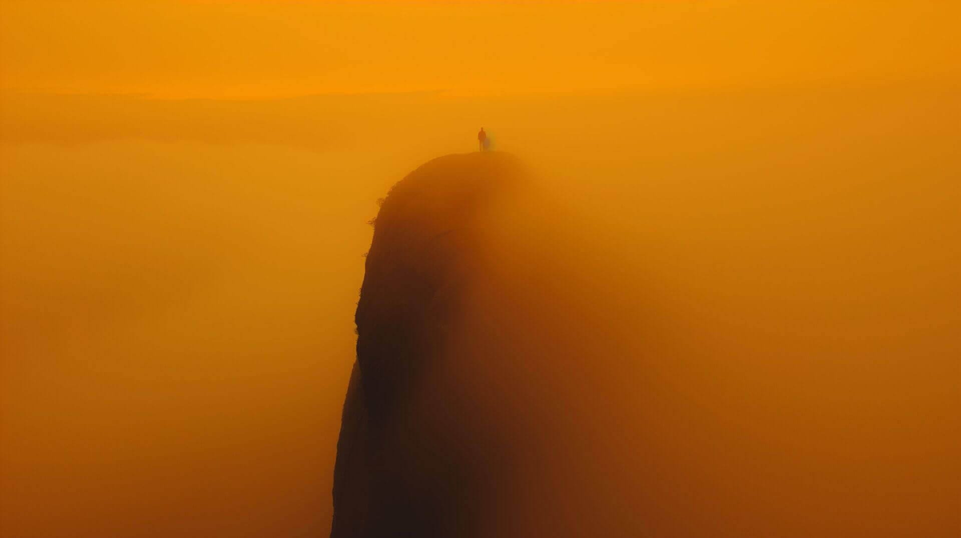 Persoon staat op een hoge bergtop omgeven door een dikke mist, die een dramatisch oranje tint heeft door de zonsondergang.