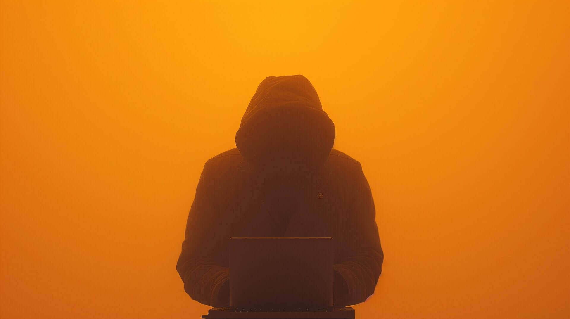 Een persoon in een donkere hoodie typt op een laptop, omgeven door een dikke oranje mist. Het gezicht van de persoon is niet zichtbaar.